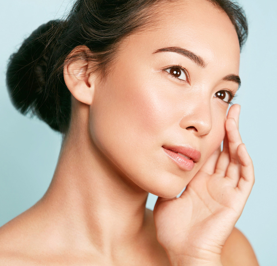 Beautiful asian girl model with natural makeup touching glowing hydrated skin on blue background closeup